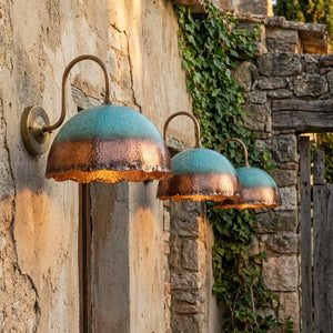 Three rustic outdoor wall sconces with hammered copper and teal-blue metal shades, mounted on a stone wall with ivy and a wooden gate in the background.
