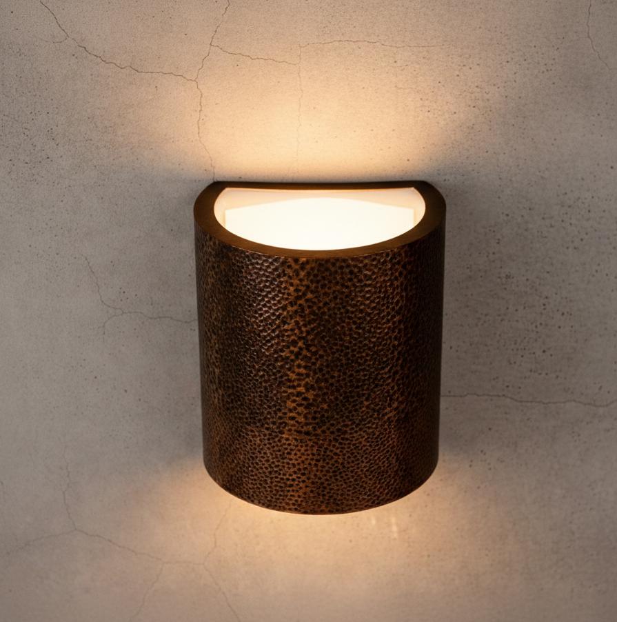 A cylindrical wall sconce with a textured, hammered metallic finish in a warm bronze-brown color, emitting a soft warm light from a semi-spherical top opening against a textured concrete wall.