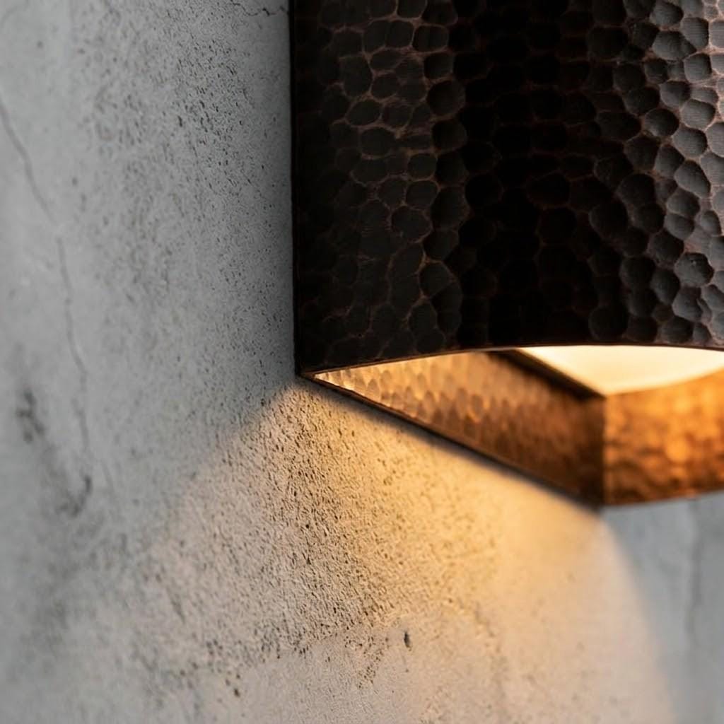 A close-up of a modern wall sconce with a hammered metal finish, mounted on a textured concrete wall and casting a warm light downward.
