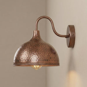 A copper-finished hammered dome wall sconce with a curved arm and round wall mount, shown lit against a neutral beige wall.
