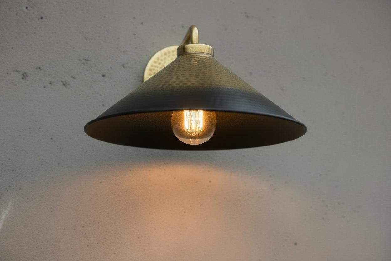 A vintage-style industrial wall sconce with a dark metal conical shade and a warm glowing light bulb, mounted on a textured light gray wall.