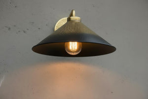 A vintage-style industrial wall sconce with a dark metal conical shade and a warm glowing light bulb, mounted on a textured light gray wall.