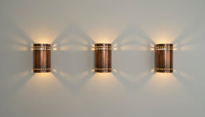 Three matching cylindrical wall sconces with a copper-toned metal finish and small cutout details, mounted on a light gray wall and casting warm light beams downward and outward.