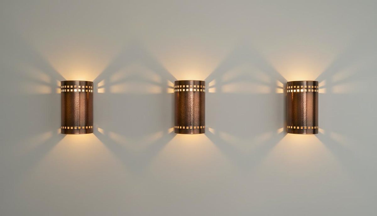 Three matching cylindrical wall sconces with a copper-toned metal finish and small cutout details, mounted on a light gray wall and casting warm light beams downward and outward.