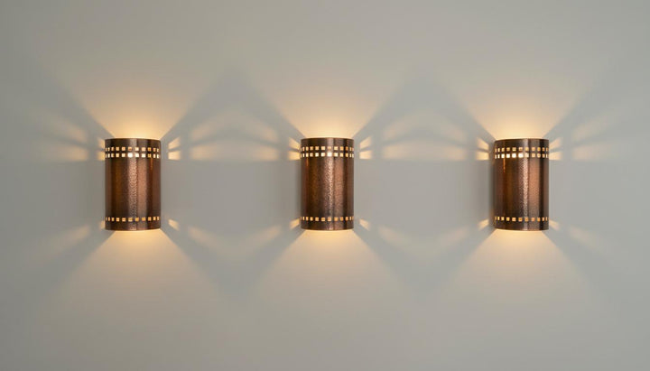 Three matching cylindrical wall sconces with a copper-toned metal finish and small cutout details, mounted on a light gray wall and casting warm light beams downward and outward.