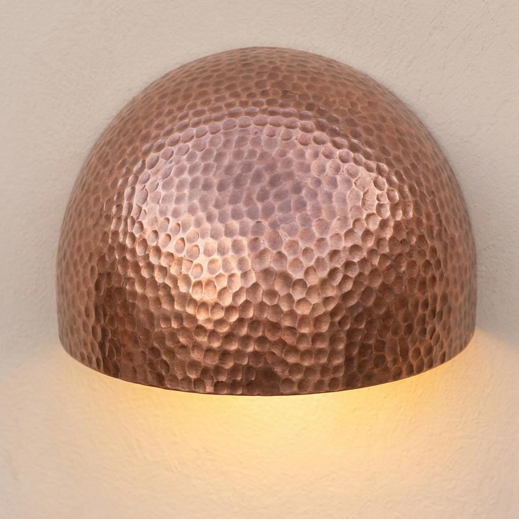 A dome-shaped hammered metal wall sconce with a warm copper finish, illuminated from within against a textured off-white wall.