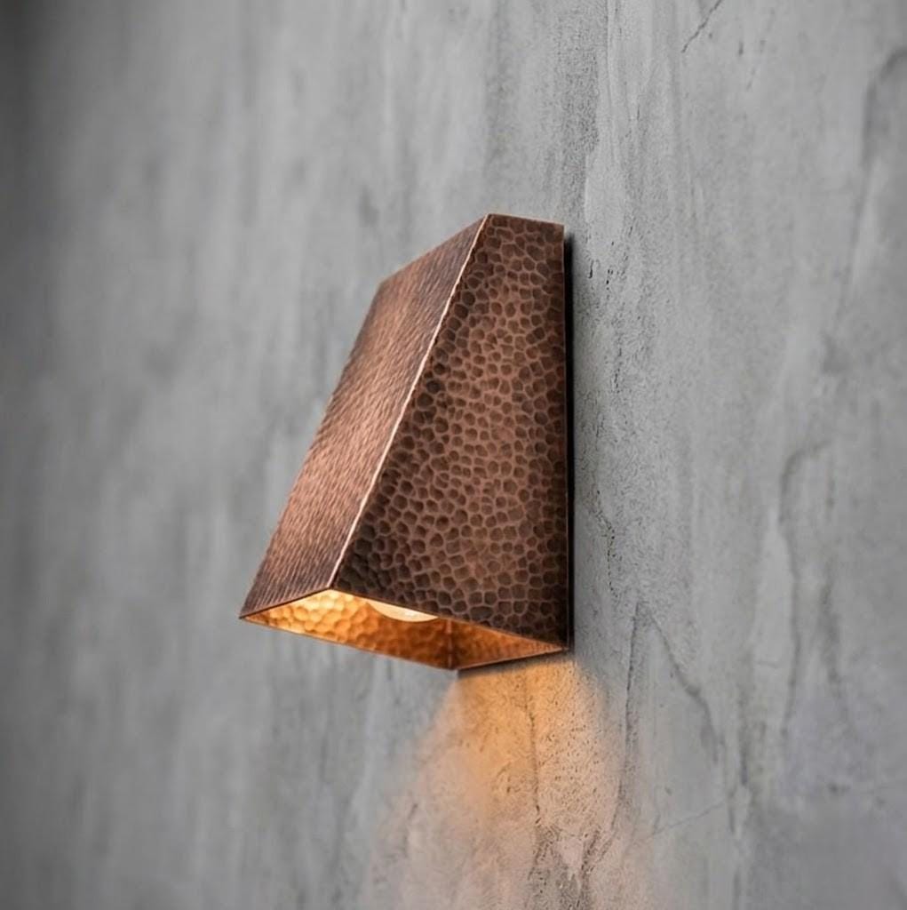 A modern geometric wall sconce with a hammered copper finish, mounted on a textured gray concrete wall and casting a warm light downward.