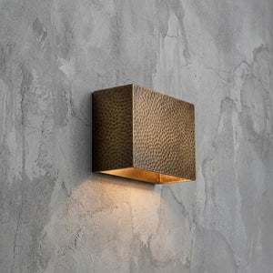 A modern square wall sconce with a hammered metallic bronze finish, mounted on a textured gray concrete wall and casting a warm light downward.