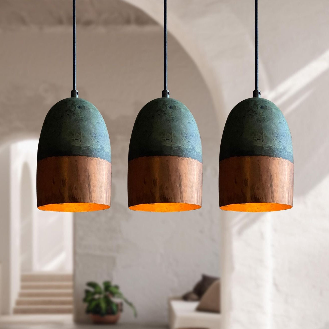 Three dome-shaped pendant lights with a textured dark green upper half and a copper-toned lower half, hanging in a softly blurred modern interior with arches and warm lighting.