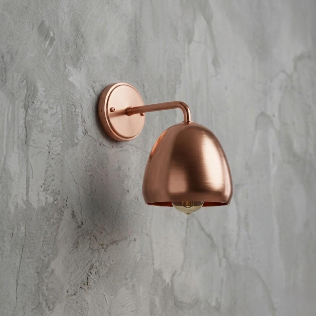 A modern wall-mounted sconce with a brushed copper dome shade and matching wall plate, mounted on a textured gray concrete wall.
