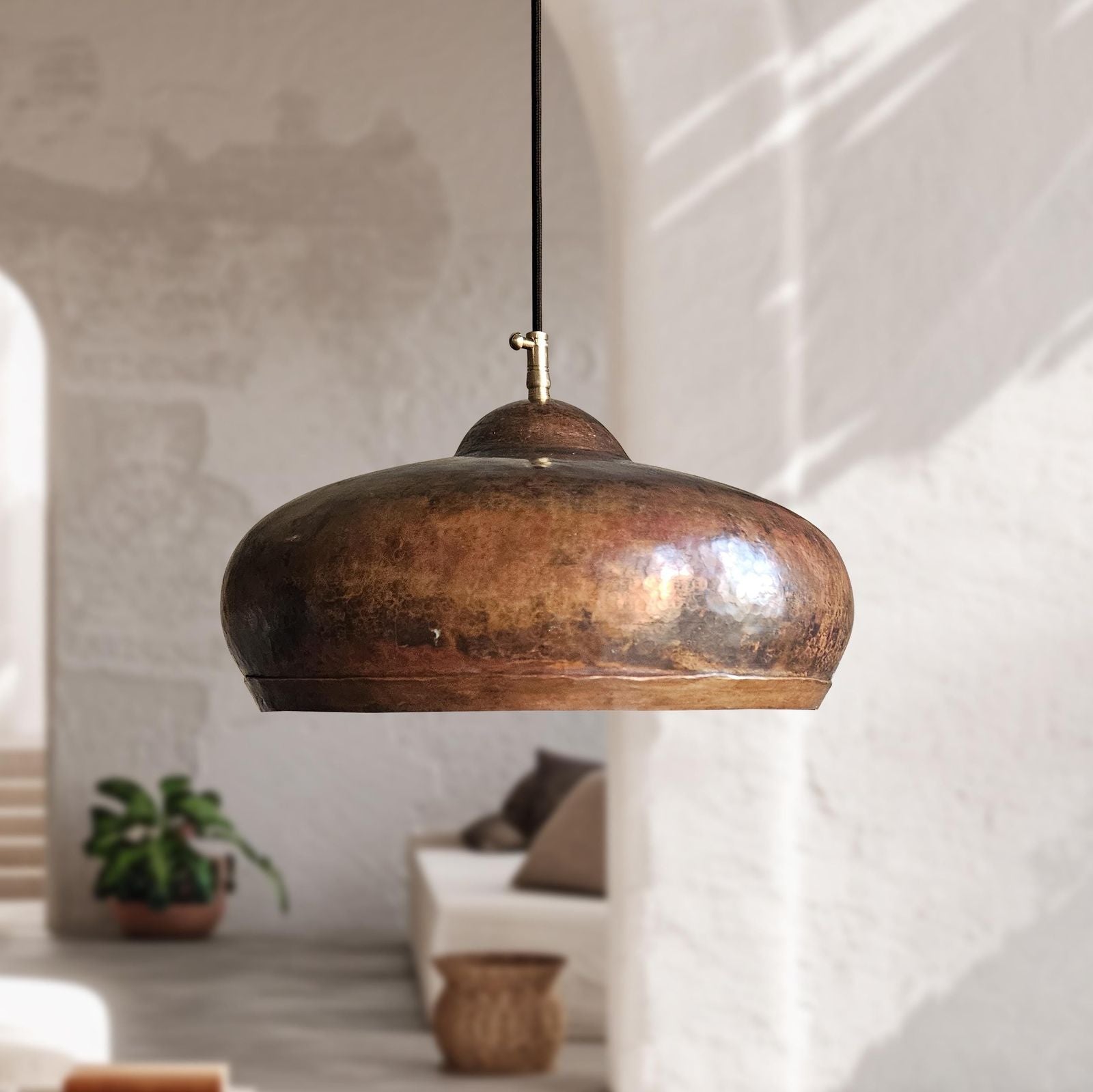 A hammered copper pendant light with a black cord, hanging in a rustic, sunlit room with white plaster walls and a blurred sofa in the background.