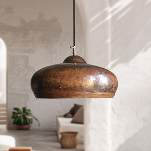 A hammered copper pendant light with a black cord, hanging in a rustic, sunlit room with white plaster walls and a blurred sofa in the background.