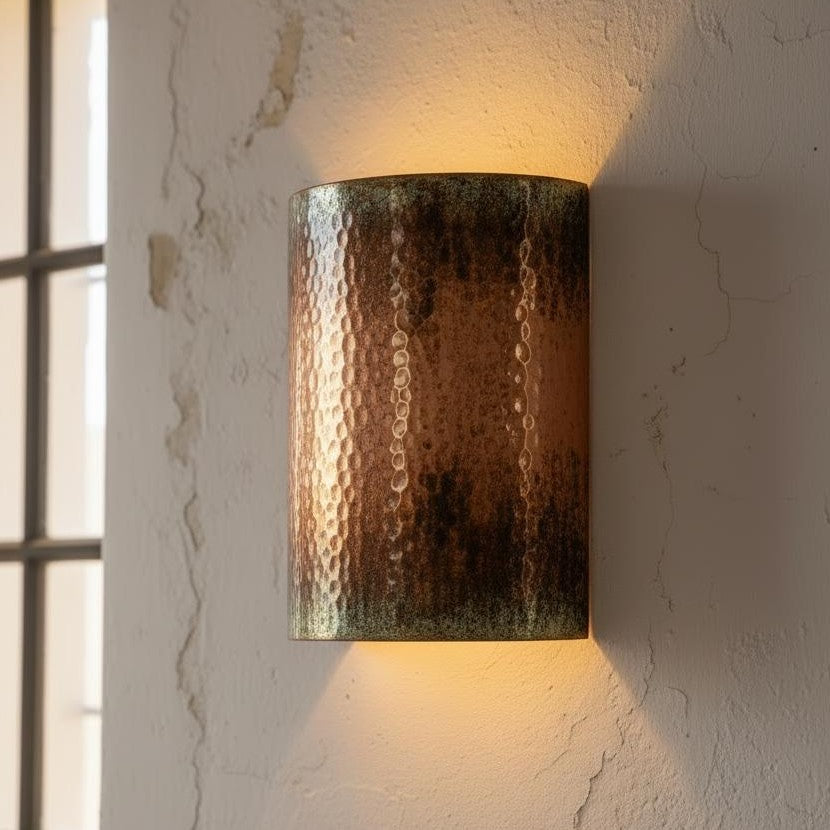 A cylindrical wall sconce with a hammered metal finish in warm bronze and copper tones, illuminated to cast a soft warm glow on a textured off-white wall.