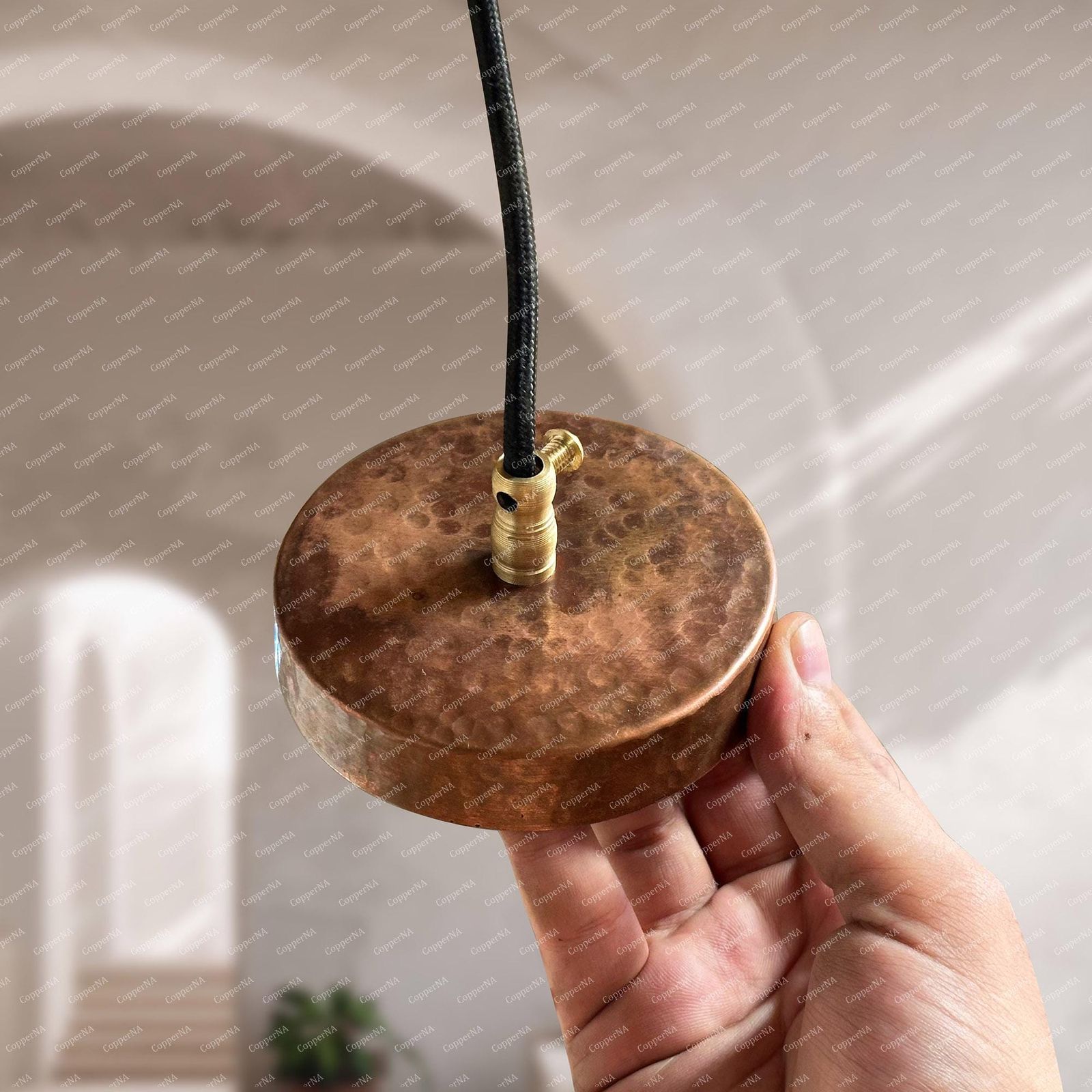 A hammered copper round pendant light base with a black cord and brass connector, held by a hand against a softly blurred interior background.