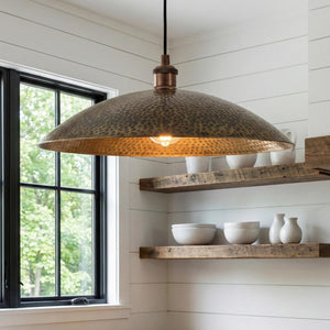 Hammered metal pendant light hanging in a kitchen with wooden shelves and windows.