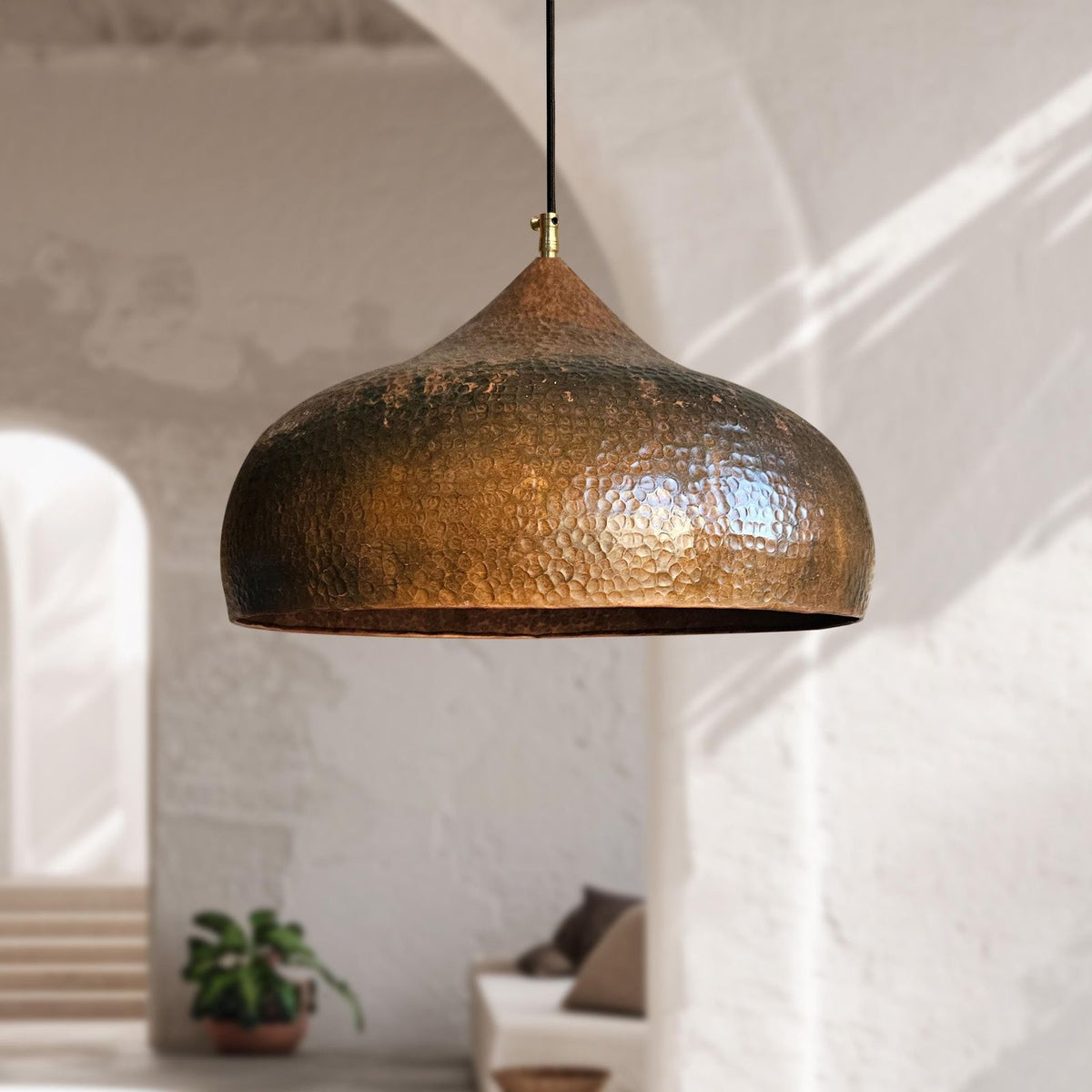 A hammered metal pendant light with a bronze finish hangs from a black cord in a bright, modern room with white walls and arches.