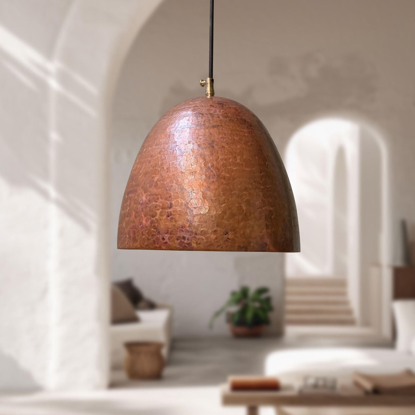 Bronze pendant lamp hanging in a softly lit room with arches and plants.
