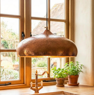 Copper pendant light hanging in a room with large windows and potted plants on a windowsill.