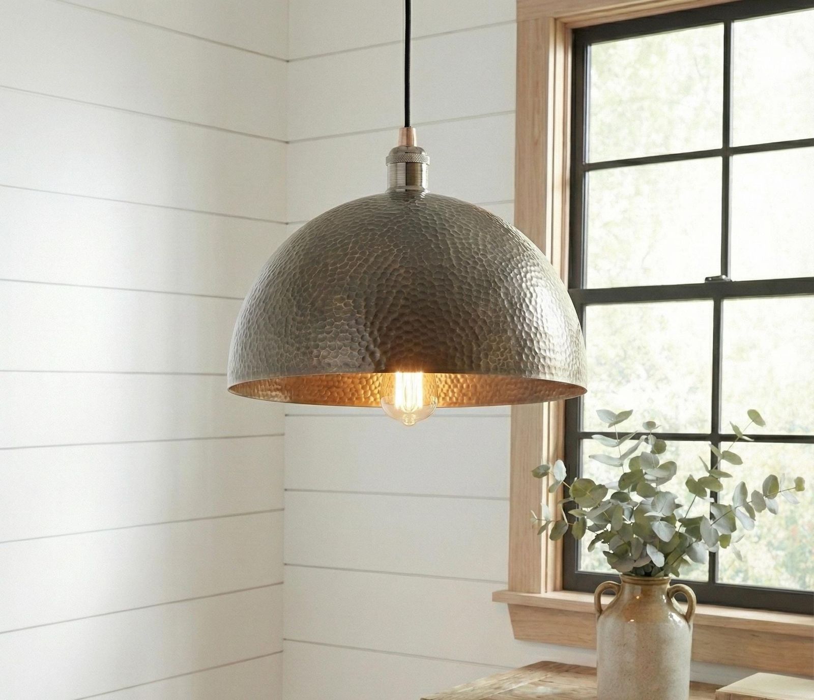 Hammered metal pendant light hanging in a room with a window and plant.