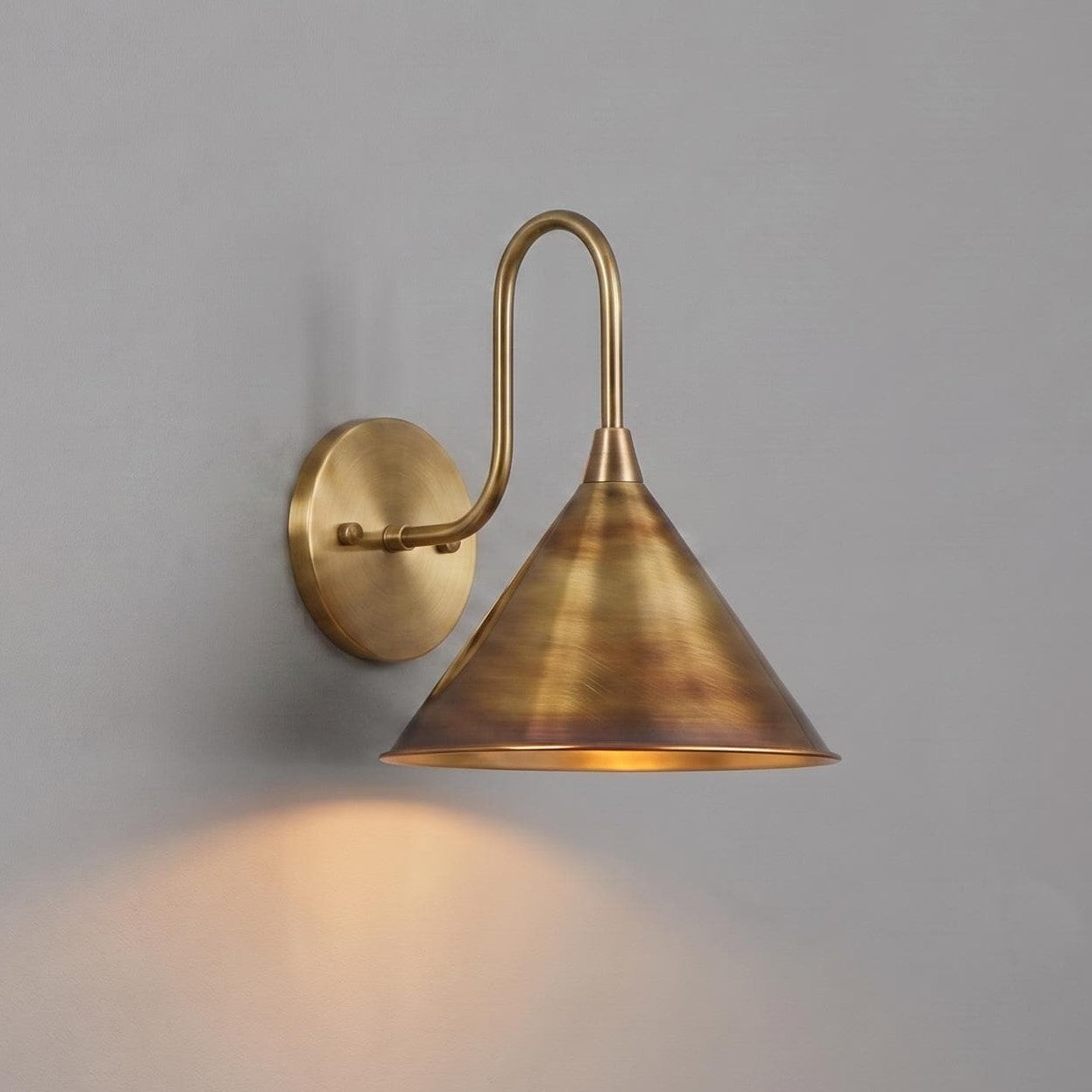 A wall-mounted sconce with a warm brass or bronze finish, featuring a circular backplate, a curved arm, and a tapered cone shade, shown lit against a plain gray wall.