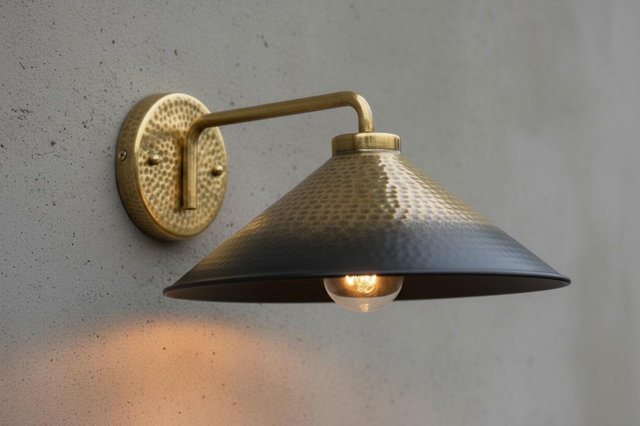 A brass wall sconce with a hammered metal shade and a visible glowing bulb, mounted on a textured gray wall.
