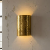 A cylindrical wall sconce with a hammered metallic gold finish, mounted on a textured off-white wall and casting a warm yellow light downward and upward.
