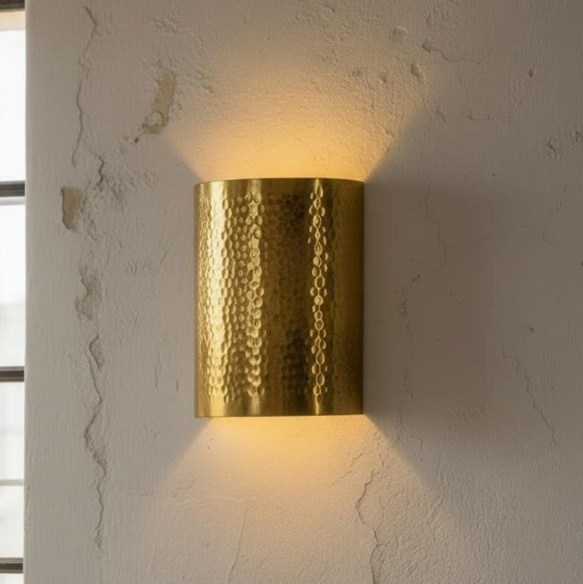 A cylindrical wall sconce with a hammered metallic gold finish, mounted on a textured off-white wall and casting a warm yellow light downward and upward.