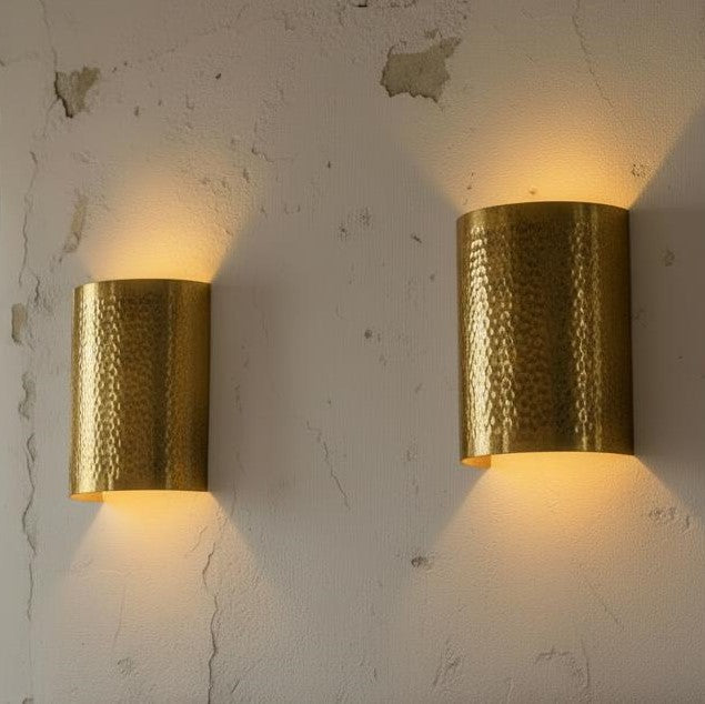 Two cylindrical wall sconces with a hammered gold finish, mounted on a textured off-white plaster wall and casting a warm yellow light.