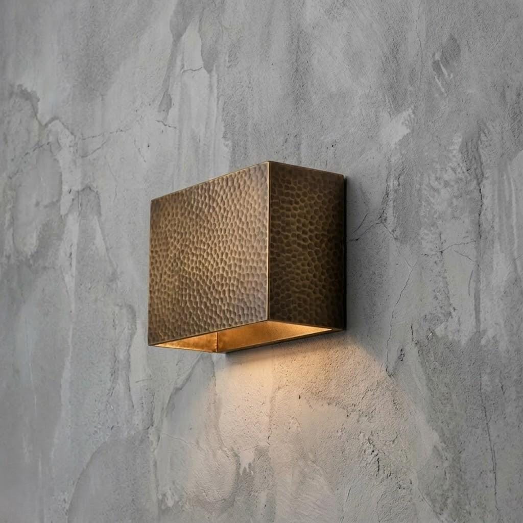 A square wall-mounted light fixture with a hammered metallic bronze finish and a warm glowing light inside, mounted on a textured gray concrete wall.