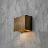 A square wall-mounted light fixture with a hammered metallic bronze finish and a warm glowing light inside, mounted on a textured gray concrete wall.