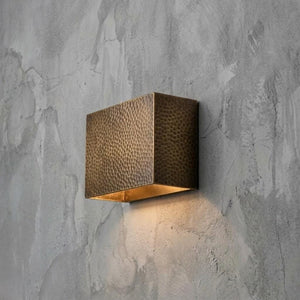 A square wall-mounted light fixture with a hammered metallic bronze finish and a warm glowing light inside, mounted on a textured gray concrete wall.
