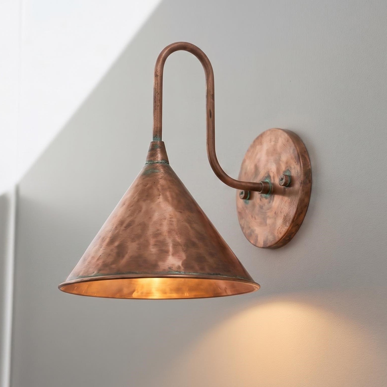 A copper wall sconce with a conical shade and curved arm, mounted on a light gray wall and casting a warm glow downward.