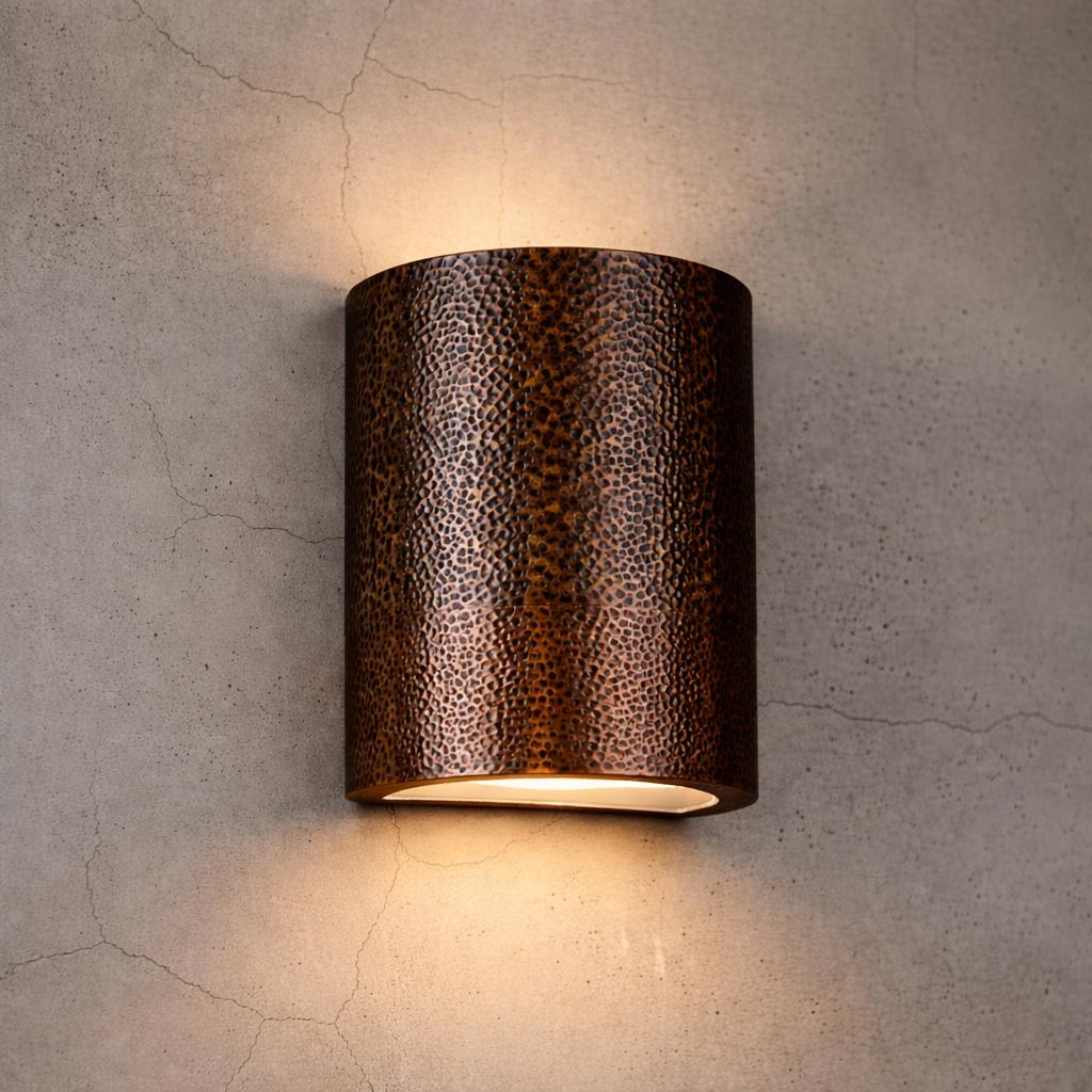 A cylindrical wall sconce with a textured, hammered metallic finish in a dark bronze-brown color, mounted on a textured concrete wall and casting a warm light downward.