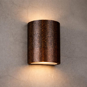 A cylindrical wall sconce with a textured, hammered metallic finish in a dark bronze-brown color, mounted on a textured concrete wall and casting a warm light downward.