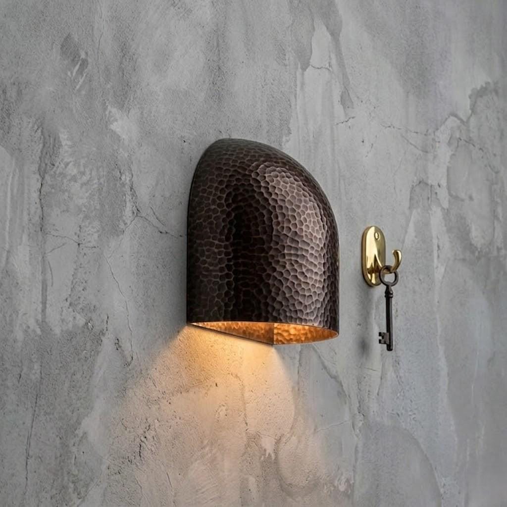 A hammered metal wall sconce in a dark bronze finish with a warm glowing light, mounted on a textured gray concrete wall next to a brass key hook.