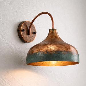 A hammered copper wall sconce with a dome-shaped shade and a warm glowing light inside, mounted on a textured off-white wall.