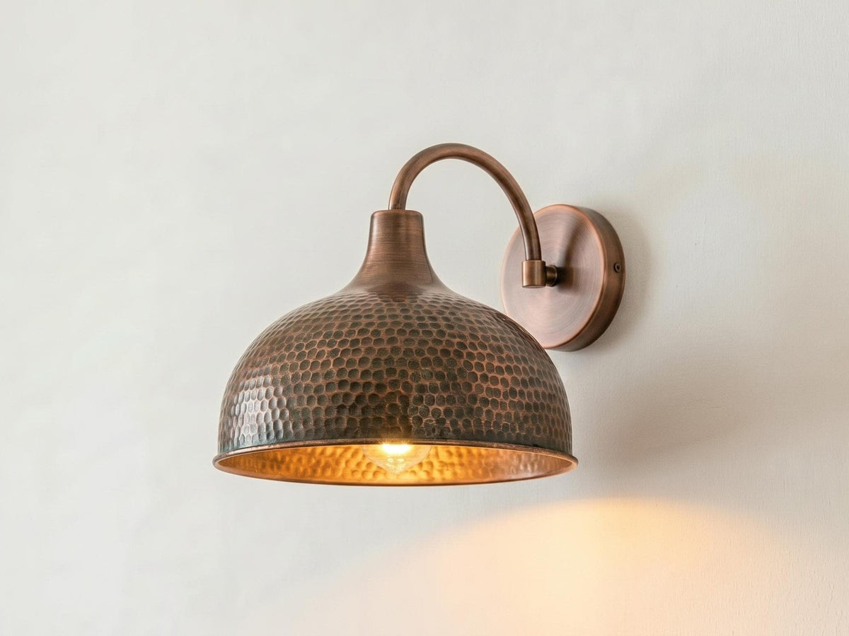 A hammered metal dome wall sconce in a warm bronze finish, mounted on a plain off-white wall and illuminated from within, casting a soft warm glow.