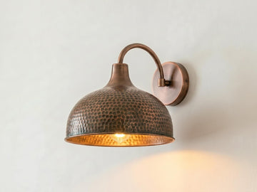 A hammered metal dome wall sconce in a warm bronze finish, mounted on a plain off-white wall and illuminated from within, casting a soft warm glow.