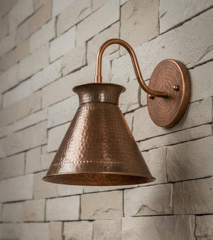 A hammered copper wall sconce with a bell-shaped shade and curved arm, mounted on a light beige stone brick wall.