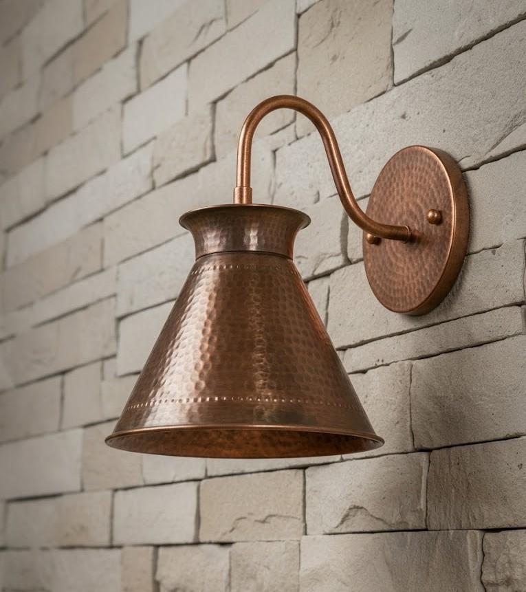 A hammered copper wall sconce with a bell-shaped shade and curved arm, mounted on a light beige stone brick wall.