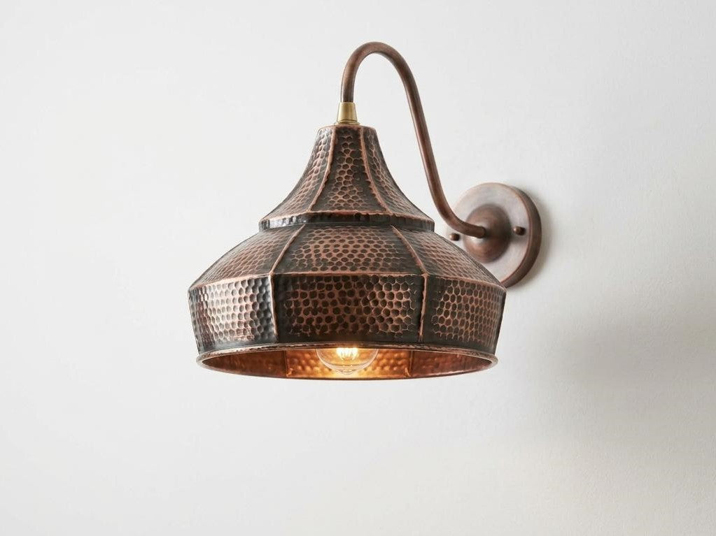 A hammered metal wall sconce with a dome-shaped shade and curved arm, finished in a warm copper/bronze tone with a glowing light inside, mounted on a plain white wall.