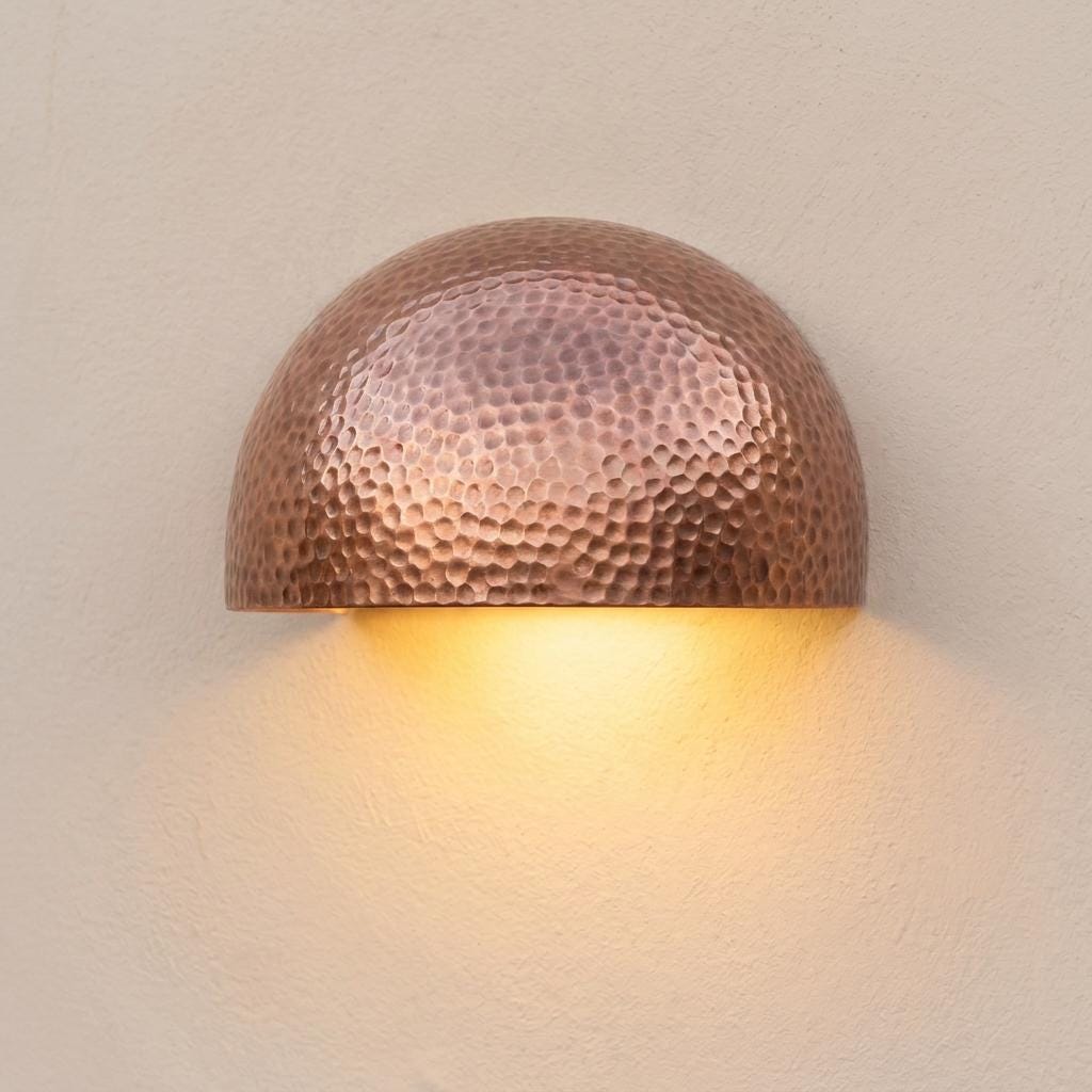 A hammered copper dome wall sconce emitting a warm yellow light against a textured beige wall.