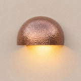 A hammered copper dome wall sconce emitting a warm yellow light against a textured beige wall.