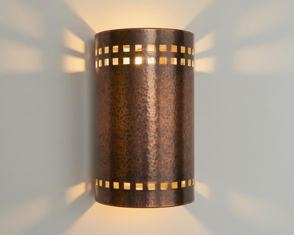 A cylindrical wall sconce with a hammered metallic finish, featuring rectangular cutouts that cast warm light onto the wall behind it.