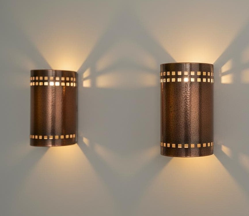 Two matching cylindrical wall sconces with a textured bronze finish and square cutout details, mounted on a light gray wall and casting warm light downward.