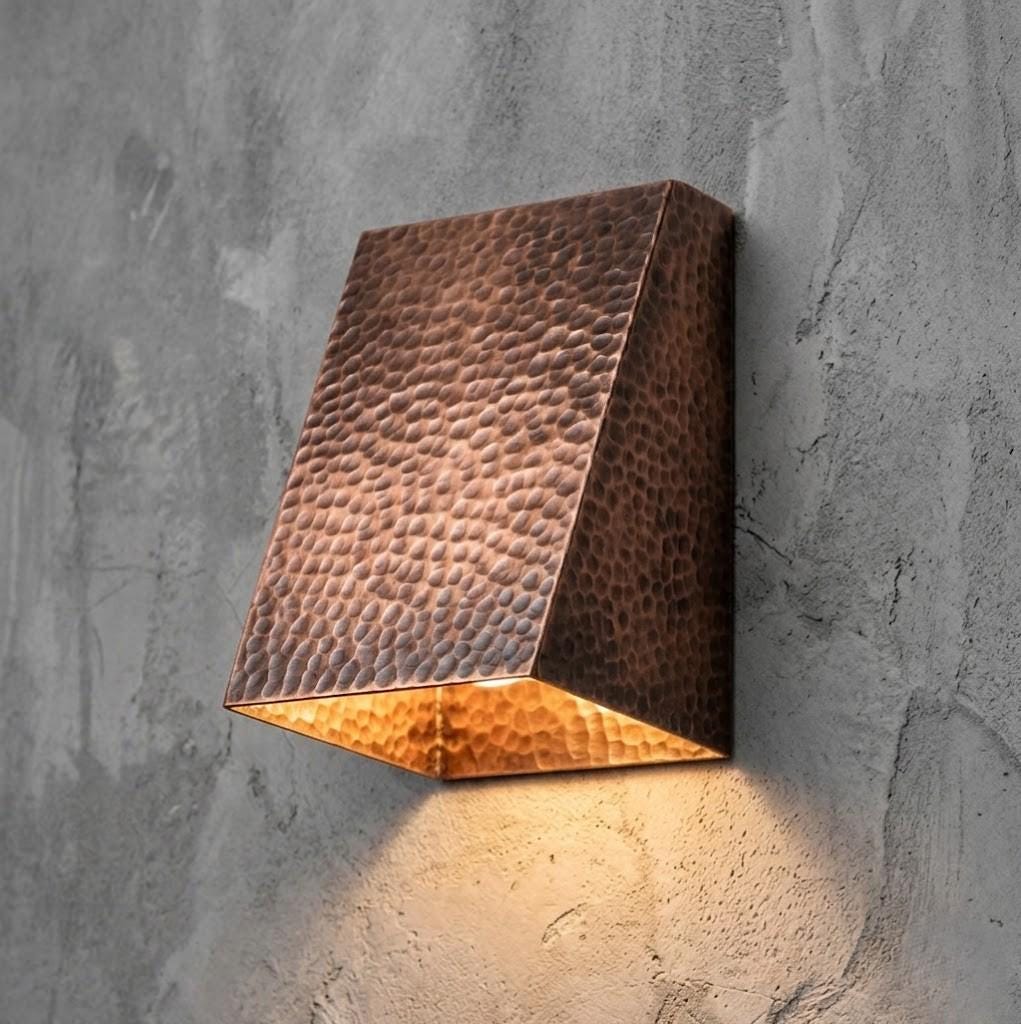 A rectangular wall sconce with a hammered copper finish, mounted on a textured gray concrete wall and casting a warm light downward.