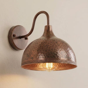 A rustic wall-mounted light fixture with a hammered copper dome shade and a curved metal arm, shown lit against a plain beige wall.