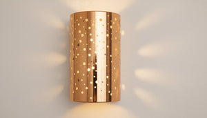 A cylindrical wall sconce with a metallic rose gold finish and laser-cut circular cutouts, casting a warm glow and dappled light pattern on a light gray wall.
