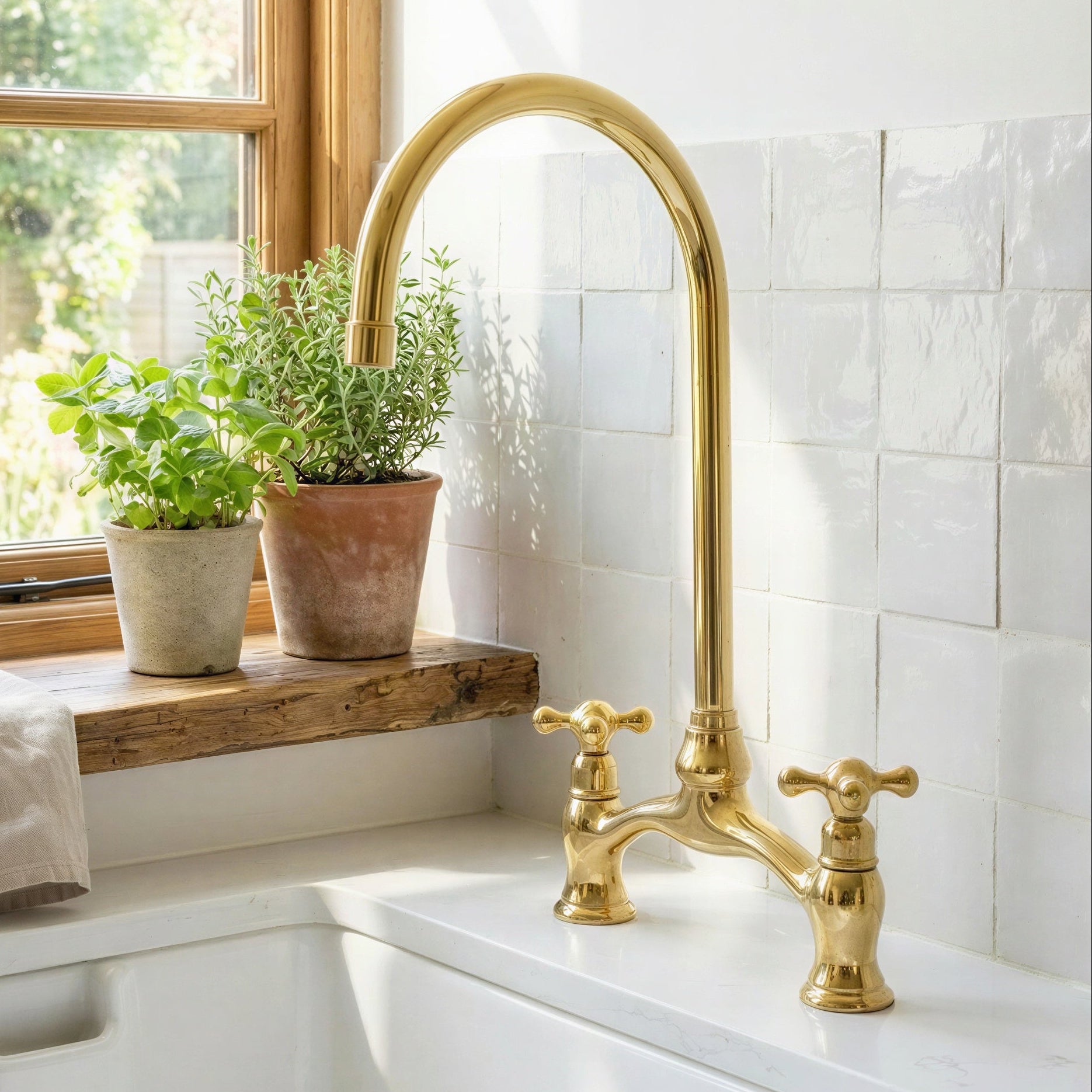 Handmade unlacquered brass bridge kitchen faucet with high-arc traditional gooseneck spout and dual cross handles mounted on white countertop next to glazed tile wall.