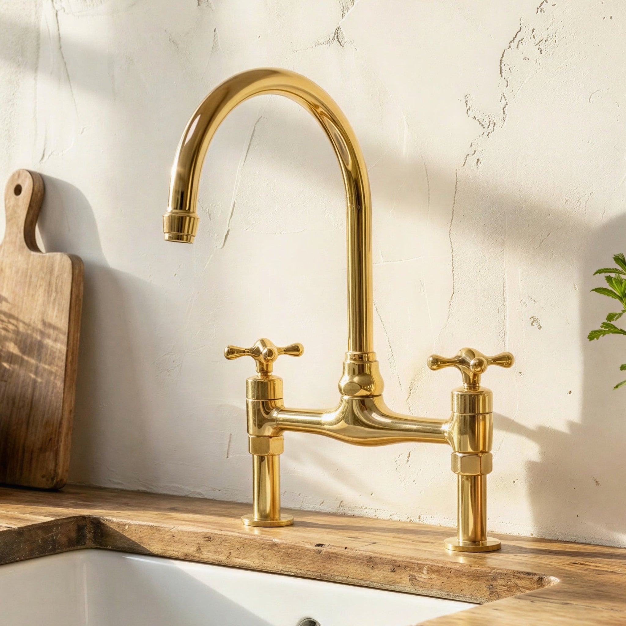 A polished brass bridge faucet with two cross handles installed over a white sink on a wooden countertop, set against a textured off-white wall with sunlight and shadow.